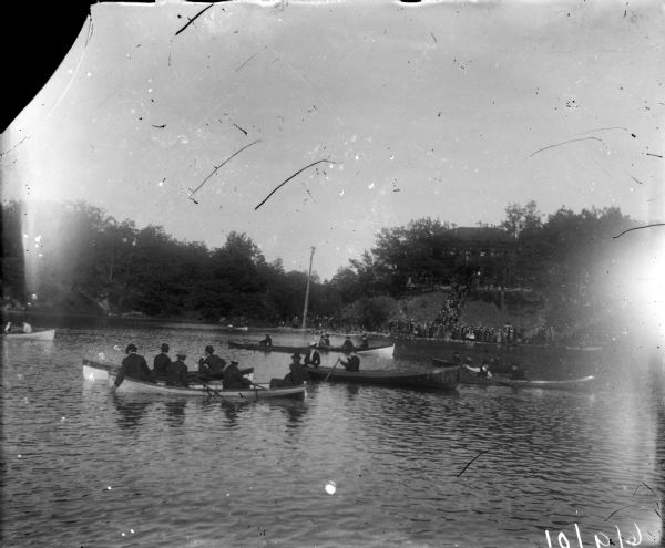 View across water of rowboats and canoes on small lake, perhaps Lake Park. On the far shoreline a large crowd is gathered, and on a hill behind them a building is visible.