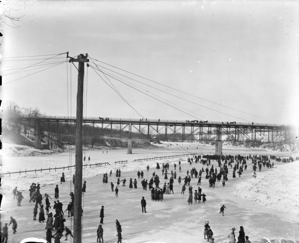 Elevated of a large group of ice skaters at River Park. Pedestrians and horse-drawn carriages can be seen on an bridge spanning the river in the background.
