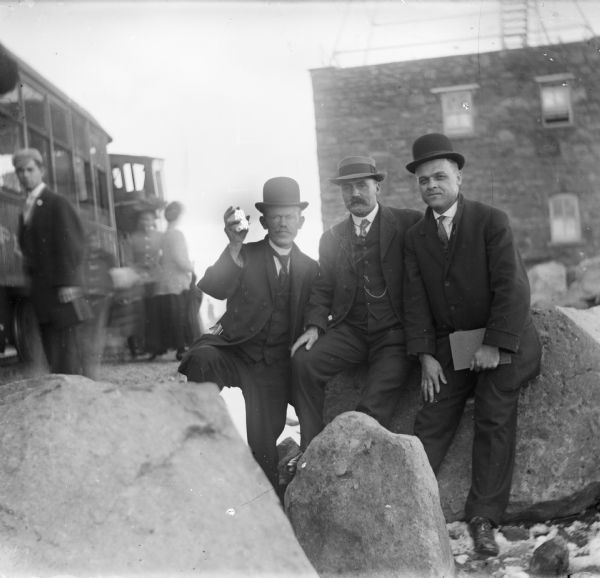 Three men posing on large rocks at Pike's Peak. Harry Dankoler is on the far left with Joe Leighton next to him and an unidentified man on the far right. A cable car and passengers are on the left in the background.