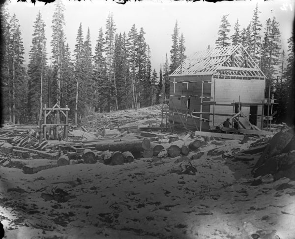 Men work on a partially finished cabin in the woods. Snow-covered tree trunks litter the clearing around the cabin. The cabin is being built in conjunction with the Leighton-Wyoming mining operation.