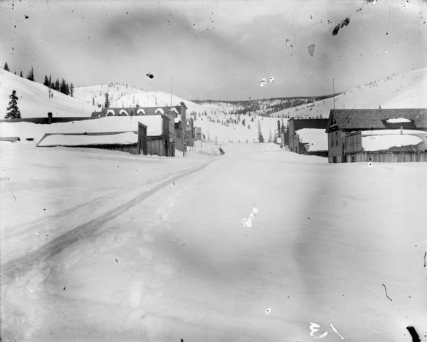 View up snowy road of a small town among low hills. A man can be seen in the far distance near some storefronts.