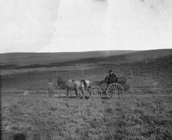 In the foreground on a hill two men stand with horse-drawn cart. In the far distance is Grand Encampment, Wyoming. The cart is headed to the Leighton-Wyoming mining camp and has 27 miles to go until it reaches its destination.