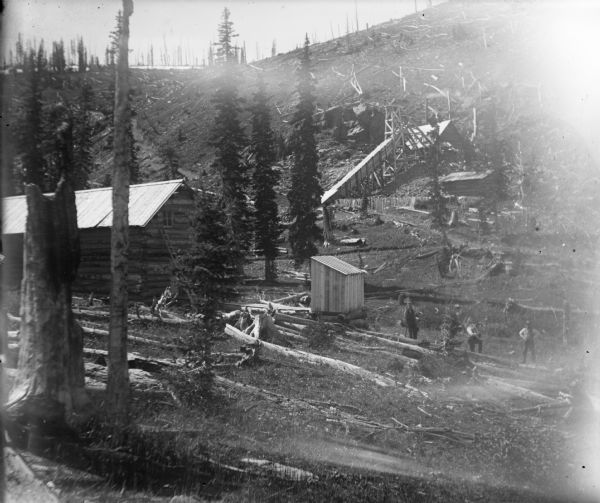 Workers in the Leighton-Wyoming mining camp. The mining operation stretches up onto a steep hill littered with fallen trees. A two-story cabin is on the left.