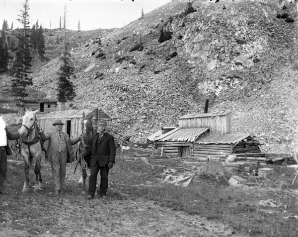 Men standing with horses in front of the Leighton-Wyoming mining camp. A steep hill rises behind them.