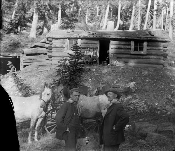Harry Dankoler, with an unidentified man, standing in front of horses and a wagon associated with the Leighton-Wyoming mining operation. Two other men sit in front of a log cabin.