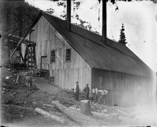 Men working at the Leighton-Wyoming mining camp stand near a large building, possibly a sawmill, related to the mining operation. A steep hill rises in the background.