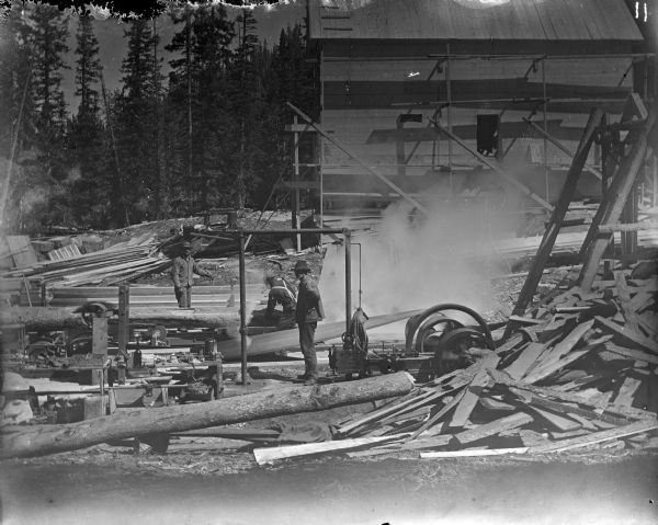 Three men working outdoors at a sawmill in the Leighton-Wyoming mining camp. In the background is a two-story industrial building.