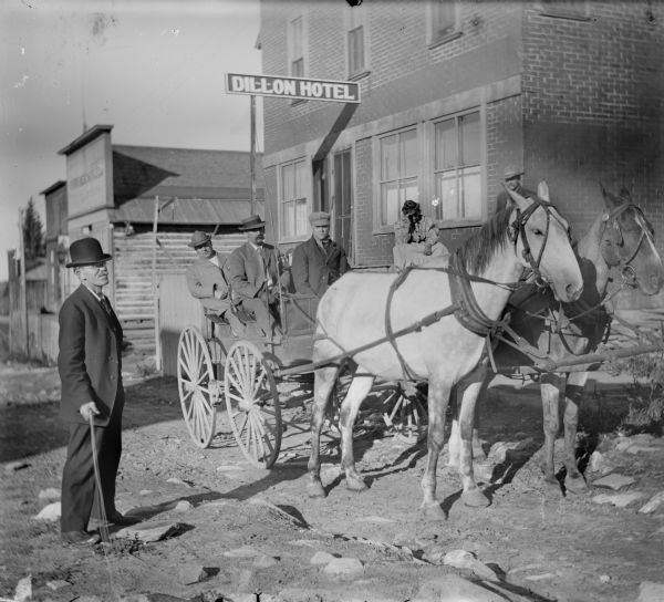 Three men are in a horse-drawn carriage in front of the Dillon Hotel where a man and woman look on from the front porch. The group is preparing to leave for the Leighton-Wyoming mines from the town of Dillon. Joe Leigton is the mustachioed man in the cart, and Harry Dankoler is on the far left holding shutter release mechanism.