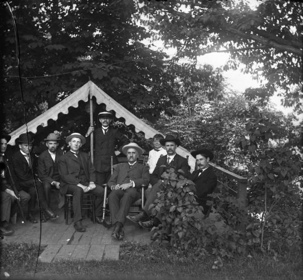 Men posing under tent on deck in backyard. Harry Dankoler is the gentleman standing directly next to the tent pole, and Syl, his son, is the young boy in the white shirt standing on in back of the group on the right.