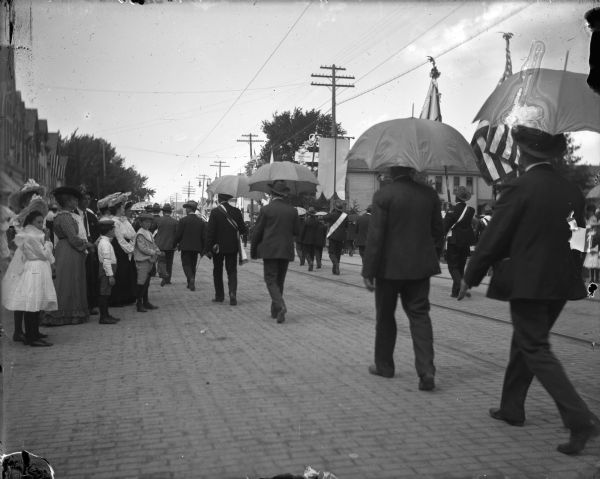 Men walking on a brick-paved street in a parade walking with umbrellas and flags while women and children watch from the sidewalk.