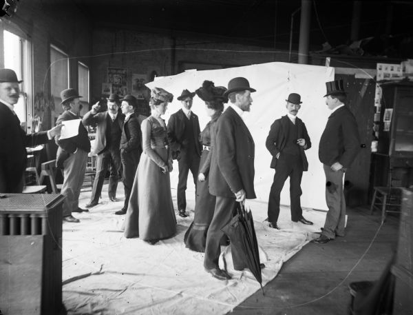 Group of men and women in a photography studio, posing in front of white backdrop. They are posed in a way that suggests a street scene. Harry Dankoler is standing on the left holding a sheet of paper.