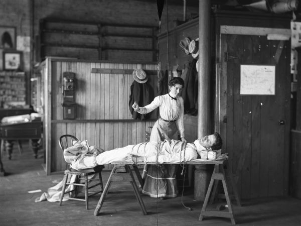 A woman standing over a man who has been tied up and laid across boards resting on two sawhorses. In the background is a telephone on a half wall, and a table or desk on the left. The image was staged at Harry Dankoler's photography studio.