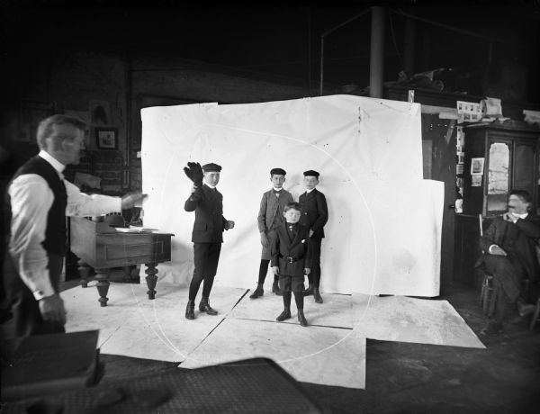 Four young boys, with Syl being the youngest, posing in Harry Dankoler's photography studio in front of a backdrop. One of the boys is holding a feather duster. Dankoler is visible giving direction in the left foreground.