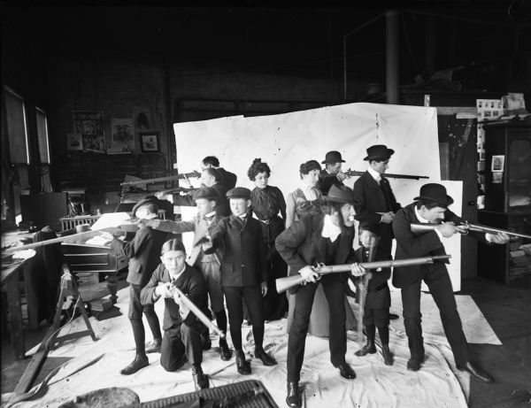 A group of men, women and children posed in Harry Dankoler's photography studio holding rifles and pistols as if they were surrounded.