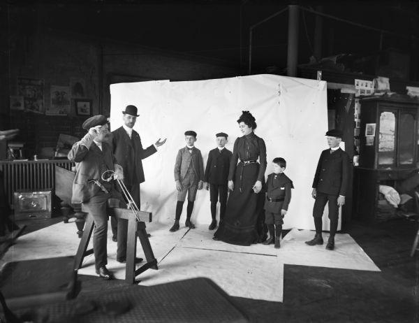 A group in front of a backdrop watches a man in the foreground ride a sawhorse as if it were a real horse. The image was staged at Harry Dankoler's photography studio.