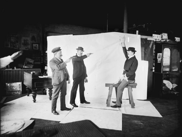 Two men are posing in front of a backdrop pointing pistols at a man who is riding a sawhorse as if it were a real horse and holding up his hand in surrender. The image was staged at Harry Dankoler's photography studio, and Dankoler's hand is visible in the left foreground.