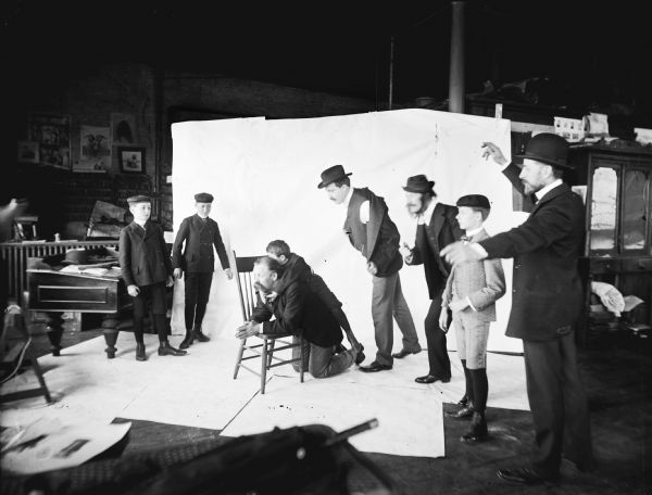 A scene staged in front of a backdrop. A man on his knees is leaning over the seat of a chair that has been turned sideways. Syl hangs on his back while three boys and three men stand around in various frozen poses. The image was staged at Harry Dankoler's photography studio.