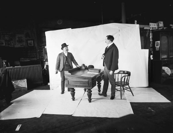 Two men posed in front of a backdrop on either side of a desk at Harry Dankoler's photography studio.