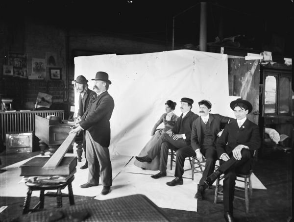 A posed photograph in front of a backdrop with two men standing to the left near a board resting on a book in the foreground. A woman and three other men are seated behind them on the right. The image was staged at Harry Dankoler's photography studio.