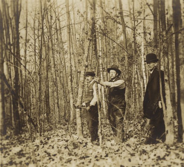 Joe Leighton, E.A. Corneille, and Harry E. Dankoler standing in a wooded area looking into the distance. Joe Leighton is holding a hatchet and E.A. Corneille appears to be holding a pipe.