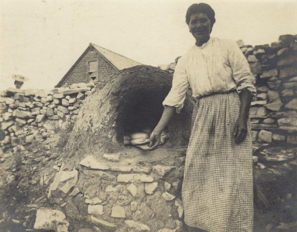 A woman adjusting bread made in a clay oven built into a stone wall. In the background is a roof of a house or shed, and a woman is looking over the stone wall.