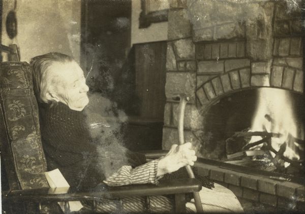 An elderly woman, most likely Dankoler's mother, sitting in a chair before a fireplace wearing a shawl. She is holding a cane in her right hand.