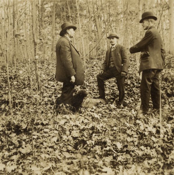 Joe Leighton, E.A. Corneille, and Harry E. Dankoler standing in a wooded area with a small dog. Dankoler is holding the holding the shutter release mechanism behind his back.