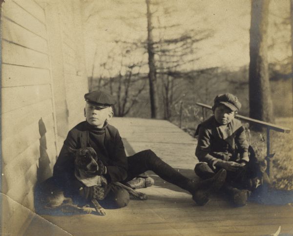 Syl, on the right, with another boy on the left holding a dog, sit on a deck outside the exterior of a building. There is a wooded yard behind them.