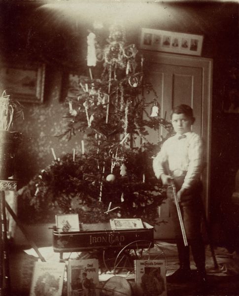 Syl standing with a rifle next to a Christmas tree surrounded by presents, including numerous books and an "Iron Clad" wagon.