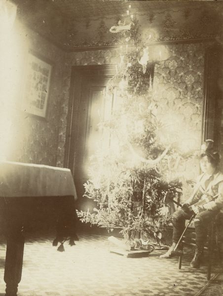 A young Syl wearing a hat, or headress, and is sitting next to a Christmas tree holding a toy in his lap. It's presumably Christmas Eve since there are no presents under the tree.