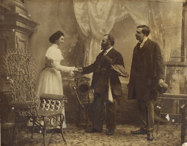 A woman who appears to be Aunt Helen shakes hands with a man in a room while another man looks on. The room appears to be a studio as it has a large painted backdrop, and an elaborate wicker chair.
