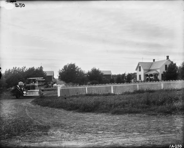 View from road of farmyard surrounded by a white picket fence. A man is riding on a horse-drawn McCormick grain binder. A small group of men are waiting inside yard near the barn. Three woman are standing on the porch of a farmhouse on the right. Several small outbuildings are in the yard.
