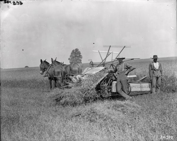 Two men posing in a field near a McCormick grain binder drawn by two mules.