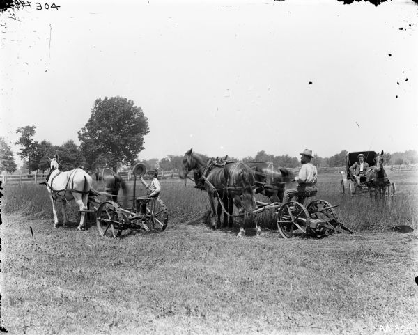 A man and a boy are each driving their own mower. Each mower is pulled by two horses.  The young boy is waving his hat. A man on the right is riding in a buggy pulled by one horse.