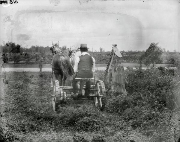Rear view of a man wearing a banded hat and riding a mower drawn by two horses. The man is lifting the sickle bar on the mower to avoid a tree stump. A road and wooden fence are in the background.