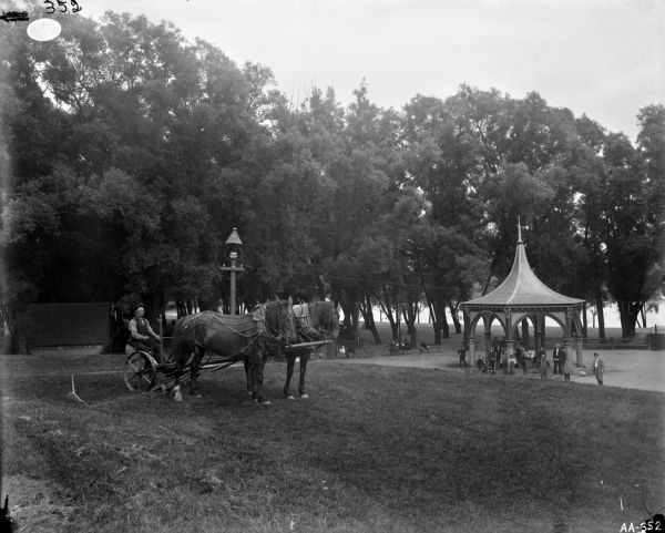 A man is sitting on a mower drawn by two horses in a park along a body of water. A group of people, including men, woman, and children, are near a band pavilion. There are park benches and bicycles in the background near large trees. A small building on the left has the words "BAGGAGE CHECKED" on the side.