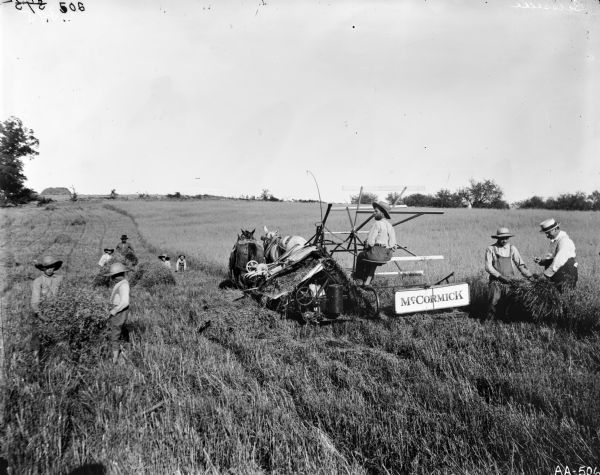 Several farmers and farm laborers, including a handful of children, at work harvesting grain with a McCormick grain binder in a field.