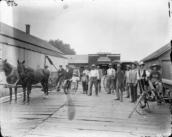 A group of farmers and young children posing on a wooden platform or boardwalk in front of a farm implement dealership building, along with a horse-drawn mower and buggy. The building has signs for Canton and P&O plows, Keystone implements, Thomas rakes, Baker wire, Morrison plows and cultivators, Charter oak wagons, and Orchard City wagons.