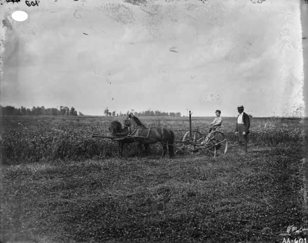 Young boy on a horse-drawn mower in a field. A man is standing nearby.