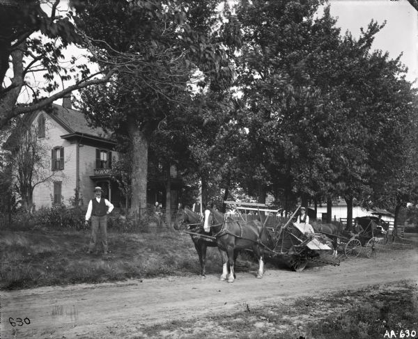 Man standing in yard in front of house and fence. There is a woman in the background sitting on the porch. A young man is driving a horse-drawn grain binder on the road in the center of the image, with two other men behind him on the edge of the road in horse-drawn carriages.