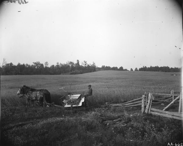 Slightly elevated view of a bearded man driving a horse-drawn McCormick grain binder in a field near a split-rail fence. There are trees in the far background.