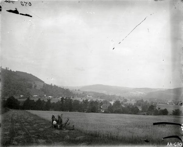 Landscape view of field, trees, and valley with farm buildings in background. A man is driving a horse-drawn reaper in a field in the foreground.