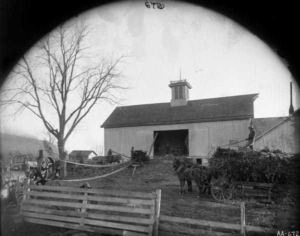 View over fence of silage cutting operation near a barn on a small hill. Several men are working in the barnyard. A horse-drawn wagon stacked with silage is being driven by a man on the right. A man and two children are standing in the open barn doorway. A stationary engine is running in the foreground.