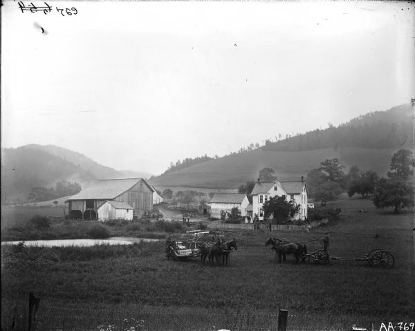 View across a field of horse-drawn grain binder with a farm in the middle distance. Men are standing in the barnyard, and a woman is standing on the lawn near the farmhouse. In the background are hills and trees in a valley. Another man appears to be standing on a horse-drawn wagon truck.