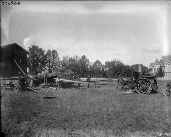 Silage operation near barn. A Westinghouse engine is powering the ensilage cutters. Men are posing near wagons. A house in the background on the right surrounded by trees and a fence.