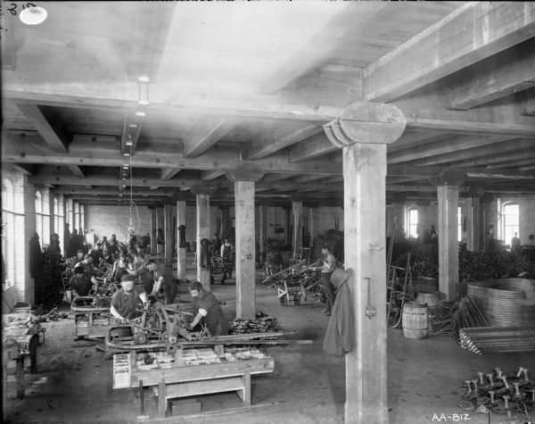 Men working at tables in the metal finishing room of the McCormick Reaper Works factory. Stacks of parts are piled on the floor, and a hat and coat are hanging from a post in the foreground.