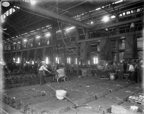 View across foundry floor of factory workers in a large room with high ceilings, vent hoods and skylights at McCormick Works. Two men in the middle ground push a wheeled cart on tracks near molds. A large crucible with long handles is sitting among molds in the foreground.