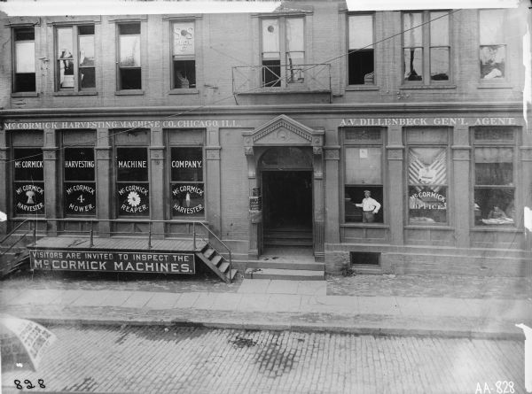 Elevated view across street of a McCormick agency(?) or dealership(?) building with man visible through a window. The windows are painted with symbols and product names. Upper line on windows read "McCORMICK HARVESTING MACHINE CO., CHICAGO, ILL." Sign hanging from platform reads "VISITORS ARE INVITED TO INSPECT THE McCORMICK MACHINES." Name painted on 2nd story window of building reads, "SS PLOW." There is an open umbrella in left foreground, which reads: "Onion Clothing Co." "A.V. DILLENBECK GEN'L AGENT," is painted above the windows to one side of the entrance.