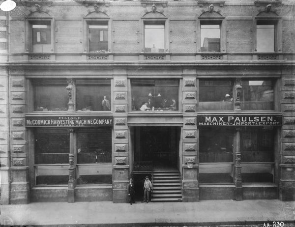 Elevated view across street of a McCormick agency(?) or dealership(?) building, possibly in Germany. Men are standing near the entrance, and other men are visible through the windows. There are two signs above first floor windows on either side of the doorway that read: "FILIALE DER McCORMICK HARVESTING MACHINE COMPANY CHICAGO U.S.A" and "MAX PAULSON. MASCHINEN-IMPORT & EXPORT."