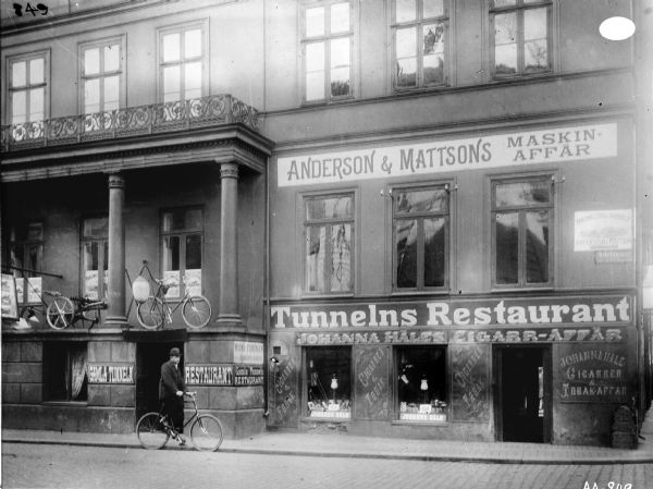 A view across Adelgatan Street of a multiple story building in Malmoe, Sweden. The store on the second floor of the building is Anderson and Mattson's machine shop and is possibly a McCormick agency or dealership. A sign on the building reads, "ANDERSON & MATTSONS MASKIN-AFFAR." On a cigar and tobacco shop signs read, "JOHANNA HALES CIGARR-AFFAR." Several other signs read, "CIGARRER & TOBAK," "Tunnelns Restaurant" and "Gamla Tunnelns RESTAURANT." A man is riding a bicycle on the street.
